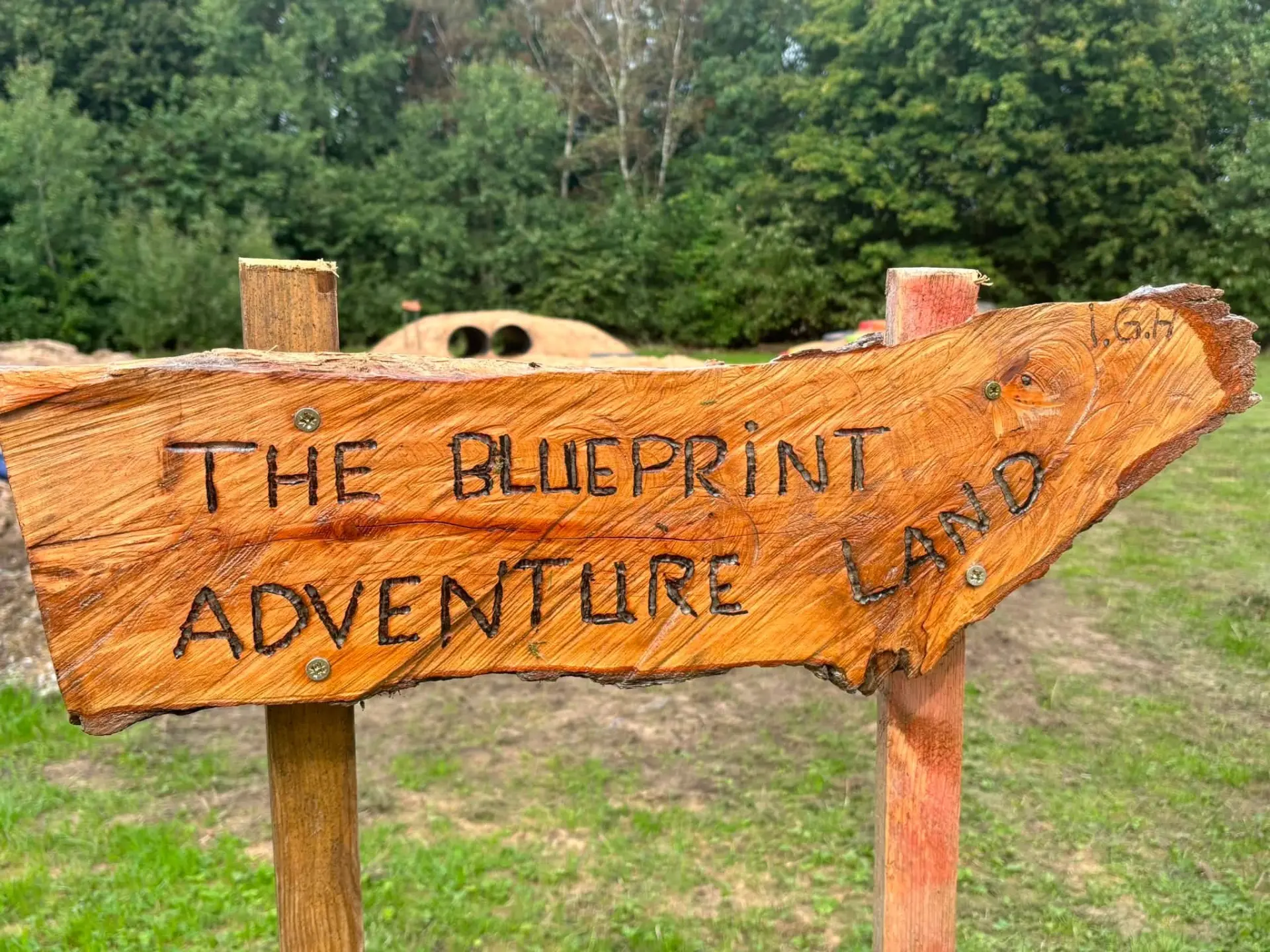 Hand-carved wooden sign reading The Blueprint Adventure Land on the school field